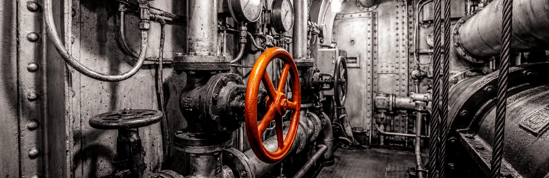 Technik, die zuverlässig im Hintergrund läuft Detail eines Maschinenraums in Schwarzweiß mit orangefarbenem Ventilrad.